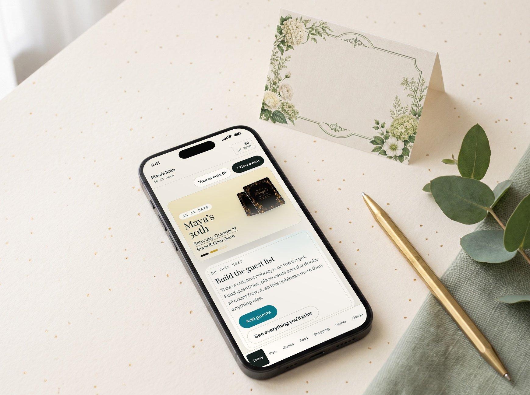 A phone showing the planner’s overview for a 30th birthday, on a cream desk beside a printed place card, a brass pen and eucalyptus